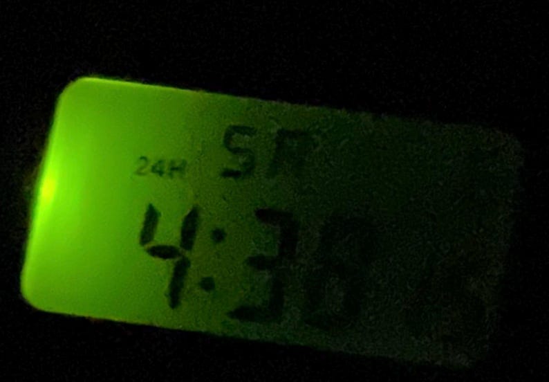 May include: A digital clock with a green illuminated display showing the time as 4:36 AM. The clock face is rectangular with rounded corners, and the background is black.