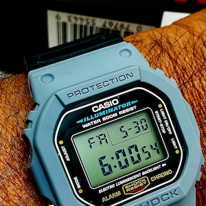 May include: A light blue Casio G-Shock digital watch with a black display showing the time and date. The watch features the text "PROTECTION", "ILLUMINATOR", and "WATER 200M RESIST". The watch also has the text "SHOCK RESIST".