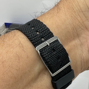 May include: A black nylon watch strap with a silver buckle. The strap is worn on a person's wrist.