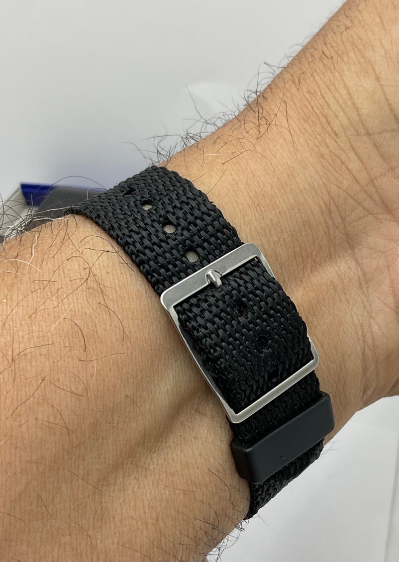 May include: A black nylon watch strap with a silver buckle. The strap is worn on a person's wrist.