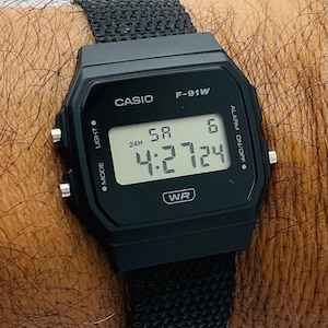 May include: Black Casio F-91W digital watch with a black fabric band. The watch face displays the time as 4:27:24. The watch has buttons for mode, light, and alarm functions. The watch is water resistant.