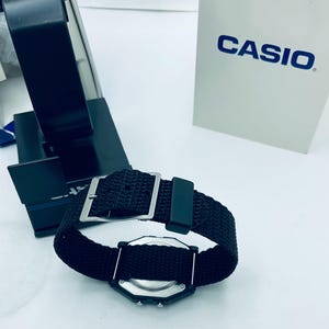 May include: A black digital watch with a black nylon strap and a silver buckle. The watch face is black with silver accents. The watch is displayed next to a white box with the word "CASIO" printed on it.