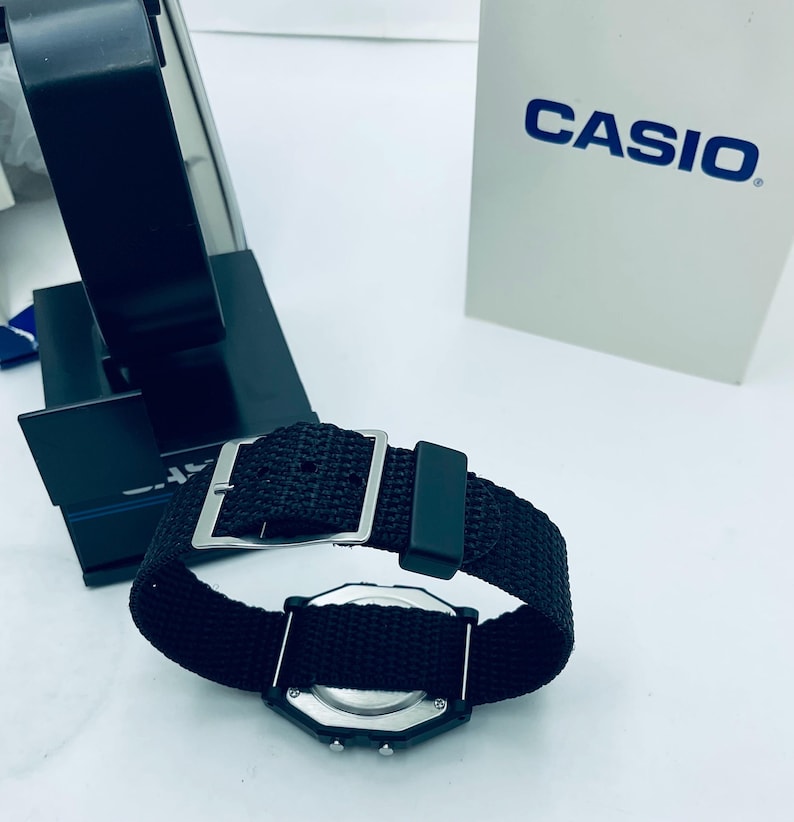 May include: A black digital watch with a black nylon strap and a silver buckle. The watch face is black with silver accents. The watch is displayed next to a white box with the word "CASIO" printed on it.