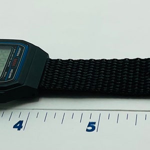 May include: A black digital watch with a black nylon strap. The watch face has a blue and white display. The watch is 8 inches long.