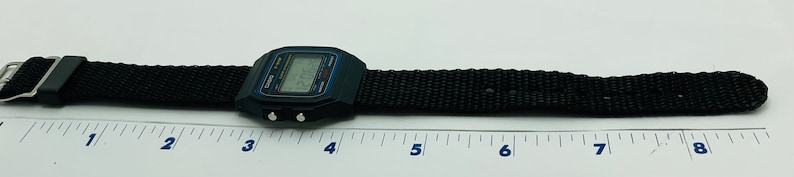 May include: A black digital watch with a black nylon strap. The watch face has a blue and white display. The watch is 8 inches long.