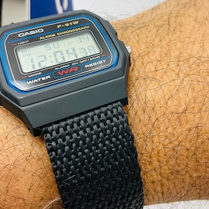 May include: A black Casio F-91W digital watch with a black nylon strap. The watch face displays the time, date, and alarm settings. The watch is water resistant.
