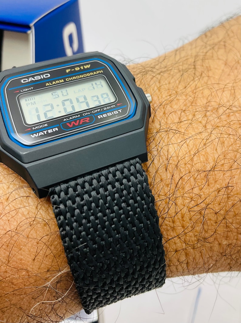 May include: A black Casio F-91W digital watch with a black nylon strap. The watch face displays the time, date, and alarm settings. The watch is water resistant.