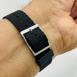 May include: Close-up of a black watch strap with a silver buckle. The strap is made of a woven material and is secured on a wrist. The watch band is a simple, yet stylish accessory.