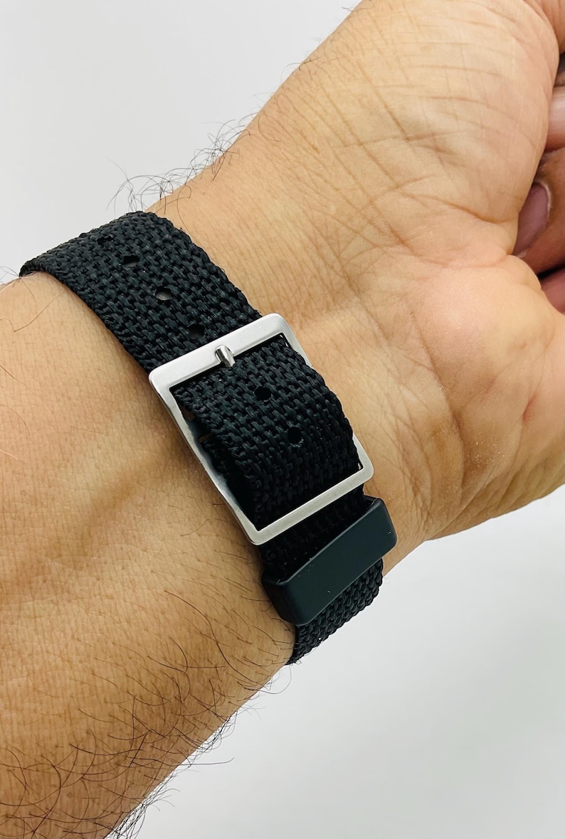 May include: Close-up of a black watch strap with a silver buckle. The strap is made of a woven material and is secured on a wrist. The watch band is a simple, yet stylish accessory.