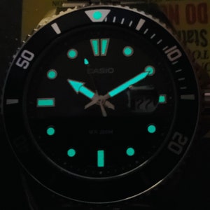 May include: A black Casio watch with a black face and silver hands. The watch has a black bezel with white markings and a white dot at the 12 o'clock position. The watch face has a green glow-in-the-dark feature. The watch face also has the text "CASIO" and "WR 200M".