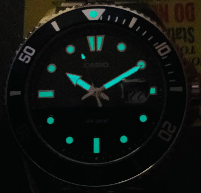 May include: A black Casio watch with a black face and silver hands. The watch has a black bezel with white markings and a white dot at the 12 o'clock position. The watch face has a green glow-in-the-dark feature. The watch face also has the text "CASIO" and "WR 200M".