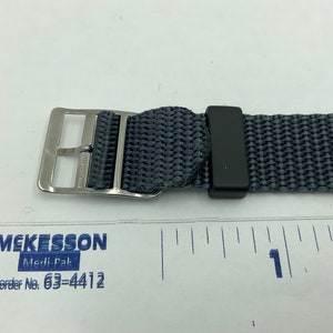 May include: A close-up of a gray nylon watch strap with a silver buckle and a black keeper. The strap is measured against a ruler, showing it is approximately 1 inch wide.
