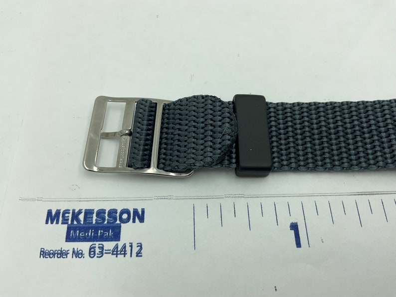 May include: A close-up of a gray nylon watch strap with a silver buckle and a black keeper. The strap is measured against a ruler, showing it is approximately 1 inch wide.