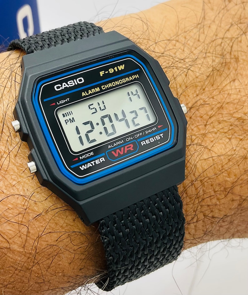 May include: A black Casio F-91W digital watch with a black woven nylon band. The watch face displays the time, date, and day of the week. The watch also has an alarm, stopwatch, and countdown timer.