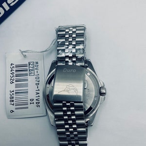 May include: A silver watch with a stainless steel strap. The watch face is black with white markings. The back of the watch has the words "Duro" and "WATER RESIST" engraved on it. The watch has a barcode sticker with the text "MDV-107D-1A1VDF 2784 DI 4549526 35887 6".