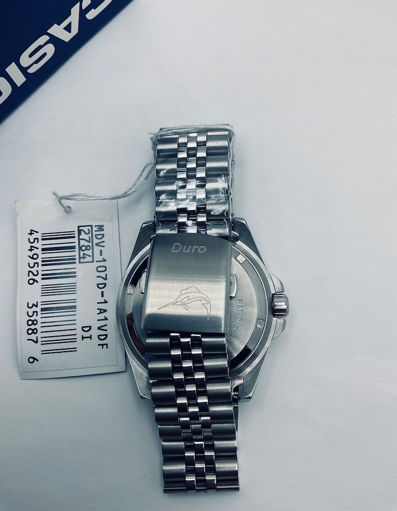 May include: A silver watch with a stainless steel strap. The watch face is black with white markings. The back of the watch has the words "Duro" and "WATER RESIST" engraved on it. The watch has a barcode sticker with the text "MDV-107D-1A1VDF 2784 DI 4549526 35887 6".