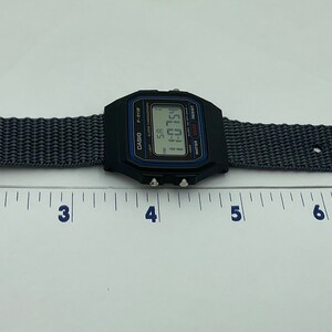 May include: A black Casio digital watch with a gray nylon strap. The watch face displays the time as 11:01. The watch is laid flat on a white surface with a ruler underneath.
