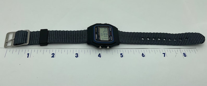 May include: A black Casio digital watch with a gray nylon strap. The watch face displays the time as 11:01. The watch is laid flat on a white surface with a ruler underneath.