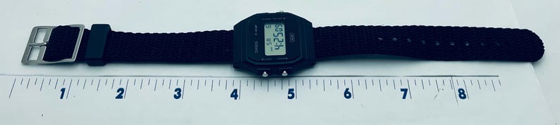 May include: Black digital watch with a black fabric band. The watch face displays the time 4:25:09. The band is approximately 8 inches long. The watch has a silver buckle.