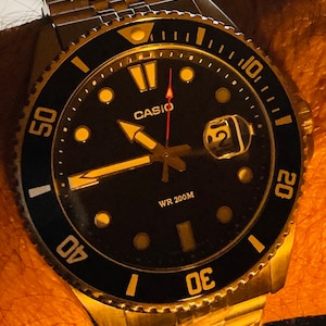 May include: A gold-toned Casio wristwatch with a black face and a silver bezel. The watch has a date window, a red second hand, and a black and gold rotating bezel. The watch is water resistant to 200 metres.