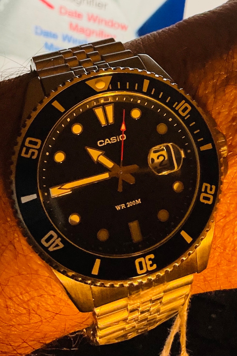 May include: A gold-toned Casio wristwatch with a black face and a silver bezel. The watch has a date window, a red second hand, and a black and gold rotating bezel. The watch is water resistant to 200 metres.