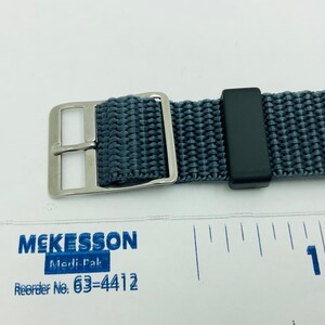 May include: A gray nylon watch strap with a silver buckle and a black keeper. The strap is about 1 inch wide.