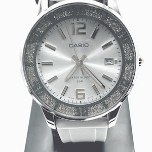 May include: A silver Casio watch with a white band. The watch face is silver with silver hands and markers. The bezel is encrusted with small, sparkling stones. The watch displays the date and is water resistant to 50 meters.