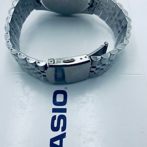 May include: A silver watch with a black face and a silver metal band. The watch is on a white surface with the word "CASIO" printed in blue.
