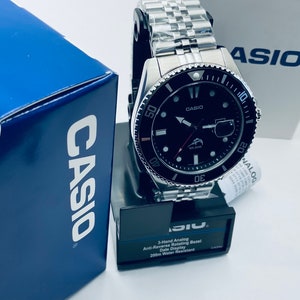 May include: A black Casio analog watch with a silver metal band and a black bezel. The watch face has a black background with silver hands and markers. The bezel has red markings and a black and silver fish logo. The watch is water resistant to 200 meters.