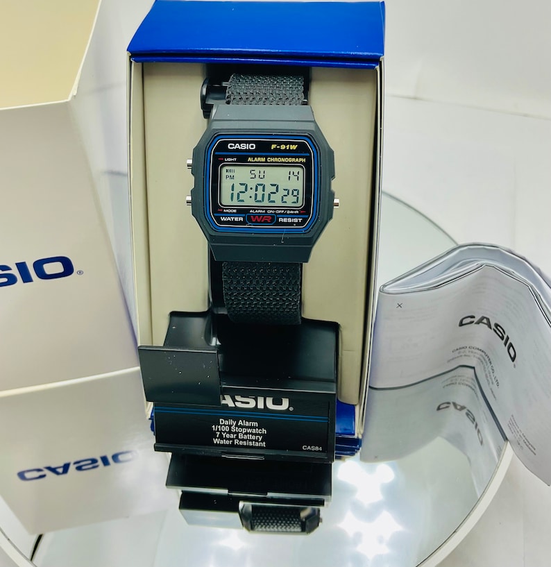 May include: A black Casio F-91W digital watch with a black fabric band. The watch face displays the time, date, and day of the week. The watch is water resistant and has a daily alarm and stopwatch.
