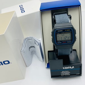 May include: A black Casio F-91W digital watch with a gray nylon strap. The watch has a blue and white display with the time, date, and alarm settings. The watch is in its original box, which is blue and white with the Casio logo.