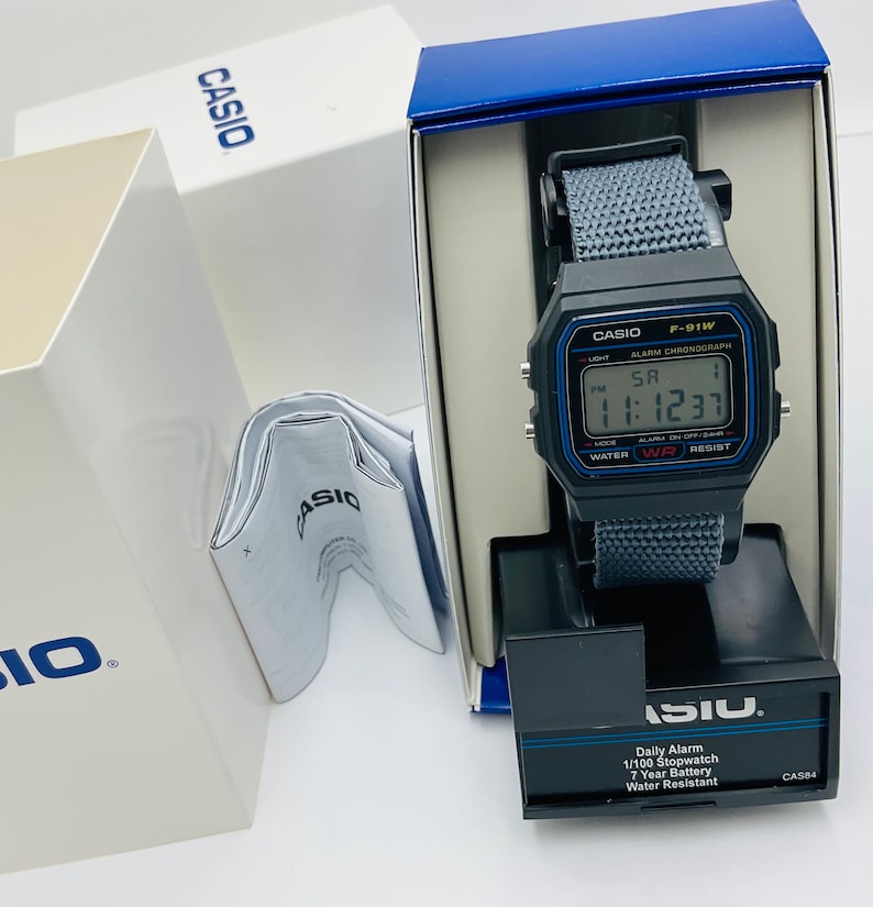 May include: A black Casio F-91W digital watch with a gray nylon strap. The watch has a blue and white display with the time, date, and alarm settings. The watch is in its original box, which is blue and white with the Casio logo.