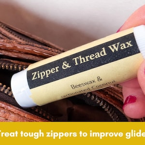 May include: A white and yellow tube of "Zipper & Thread Wax" with black text, held near a brown zipper. The label also lists "Beeswax & fractionated Coconut". The image includes the text "Treat tough zippers to improve glide."