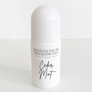 Pode incluir: Frasco de desodorizante roll-on branco com um r&oacute;tulo preto que diz "Magnesium Deodorant by Weathered Door" e "Cedar Mint" em uma fonte manuscrita.