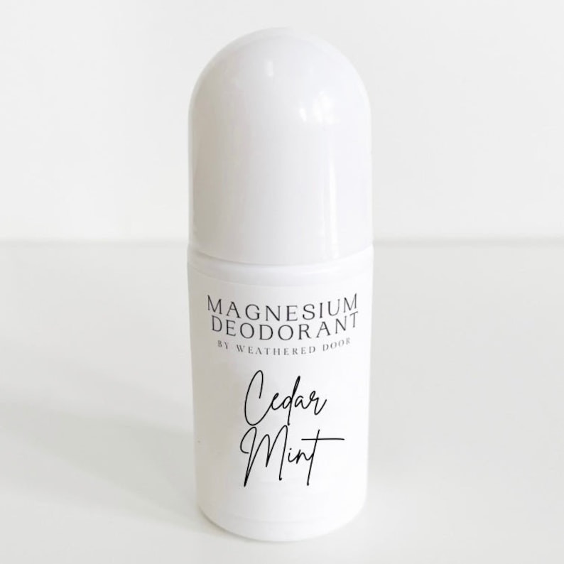Pode incluir: Frasco de desodorizante roll-on branco com um r&oacute;tulo preto que diz "Magnesium Deodorant by Weathered Door" e "Cedar Mint" em uma fonte manuscrita.