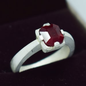 Könnte beinhalten: Ein silberner Ring mit einem roten Edelstein, der in einer quadratischen Fassung sitzt.