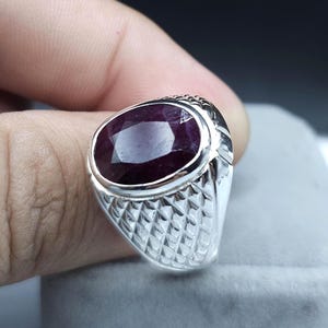 May include: A silver ring featuring a large, oval-cut, deep red gemstone. The ring has a textured, diamond-patterned band and a smooth bezel setting. The gemstone is the focal point, showcasing its rich color and clarity.