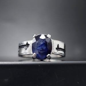 May include: A silver ring featuring a large, oval-cut, deep blue gemstone. The ring has a wide band with decorative black accents. The gemstone is set with four prongs, and the ring is displayed against a dark background.