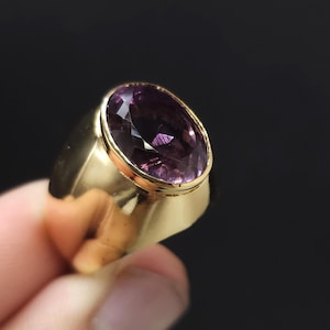 May include: A gold ring featuring a large, oval-cut purple gemstone set in a smooth, polished gold band. The gemstone displays deep violet tones with multifaceted surfaces that reflect light, while the band has a wide, solid, and reflective finish.