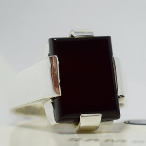 Könnte beinhalten: Ein silberner Ring mit einem schwarzen rechteckigen Edelstein. Der Ring hat ein schlichtes Design mit einer glatten Ringschiene und einer Zargenfassung für den Stein.