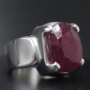 May include: A silver ring with a large, faceted, oval-shaped ruby gemstone.