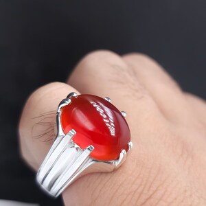 May include: A silver ring with a large, oval-shaped, red gemstone set in a claw setting. The ring has a thick band with a textured design.