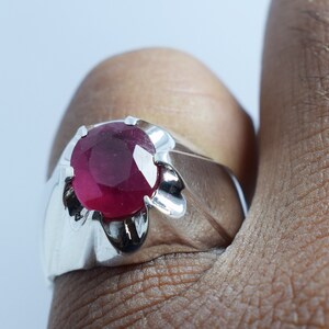 Może przedstawiać: Srebrny pierścionek z dużym, okrągłym, czerwonym rubinem osadzonym w szponach. Pierścionek ma szeroką obrączkę z fakturowanym wzorem.