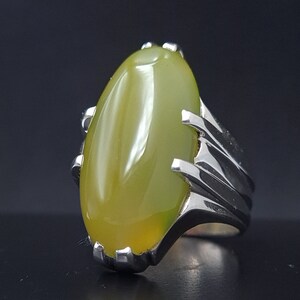 May include: A silver ring with a large, oval-shaped, light green gemstone set in a claw setting. The band of the ring is silver and has a textured design.