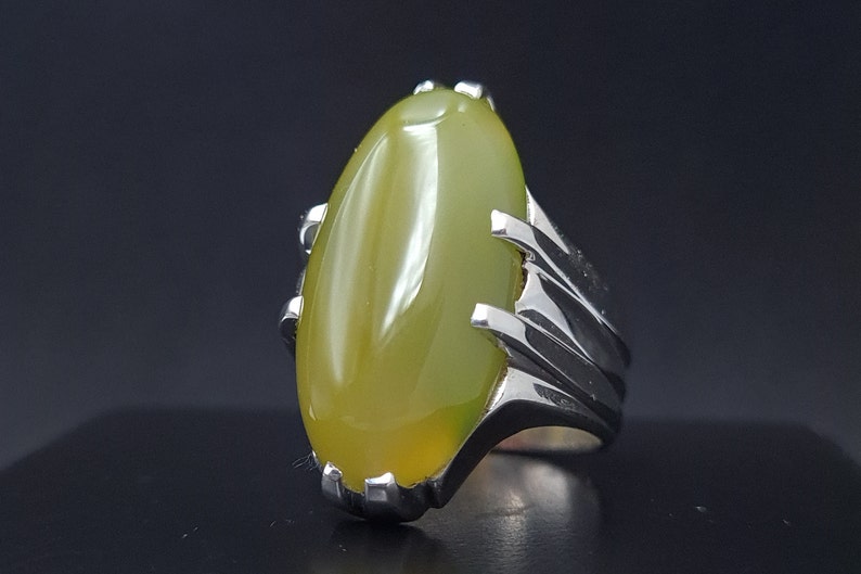 May include: A silver ring with a large, oval-shaped, light green gemstone set in a claw setting. The band of the ring is silver and has a textured design.