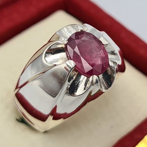 Pode incluir: Um anel de prata com uma grande gema de rubi rosa ovalada engastada no centro. O anel tem uma banda texturizada e detalhada.