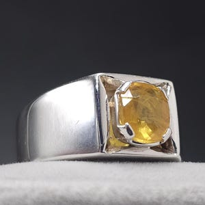May include: A silver ring with a large, square setting. The ring features a vibrant, yellow gemstone. The gemstone is a cushion cut and is securely held in place by four prongs. The ring is displayed on a white surface.