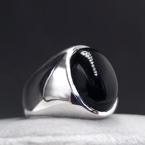 Op de afbeelding: Een zilveren ring met een grote, ovale, zwarte onyx steen. De ring heeft een gepolijste, gladde afwerking en is geplaatst op een wit oppervlak tegen een donkere achtergrond. De onyx steen is zeer reflecterend.
