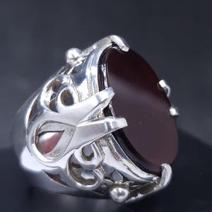 Puede incluir: Un anillo de plata con un diseño adornado y una piedra ovalada de color rojo oscuro.