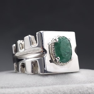 May include: A silver ring with a large, oval-cut green gemstone. The ring has a unique design with a rectangular face and a decorative side. The gemstone is set in a detailed bezel.